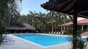A swimming pool with lounge chairs, a thatched-roof hut, and palm trees. - Hemingway By YellowKey (Guayacanes)