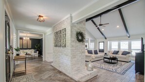 A spacious living room with a fireplace, a ceiling fan, and a large window. - Chic TX House w/ Pool and Relaxing Atmosphere (Spring)