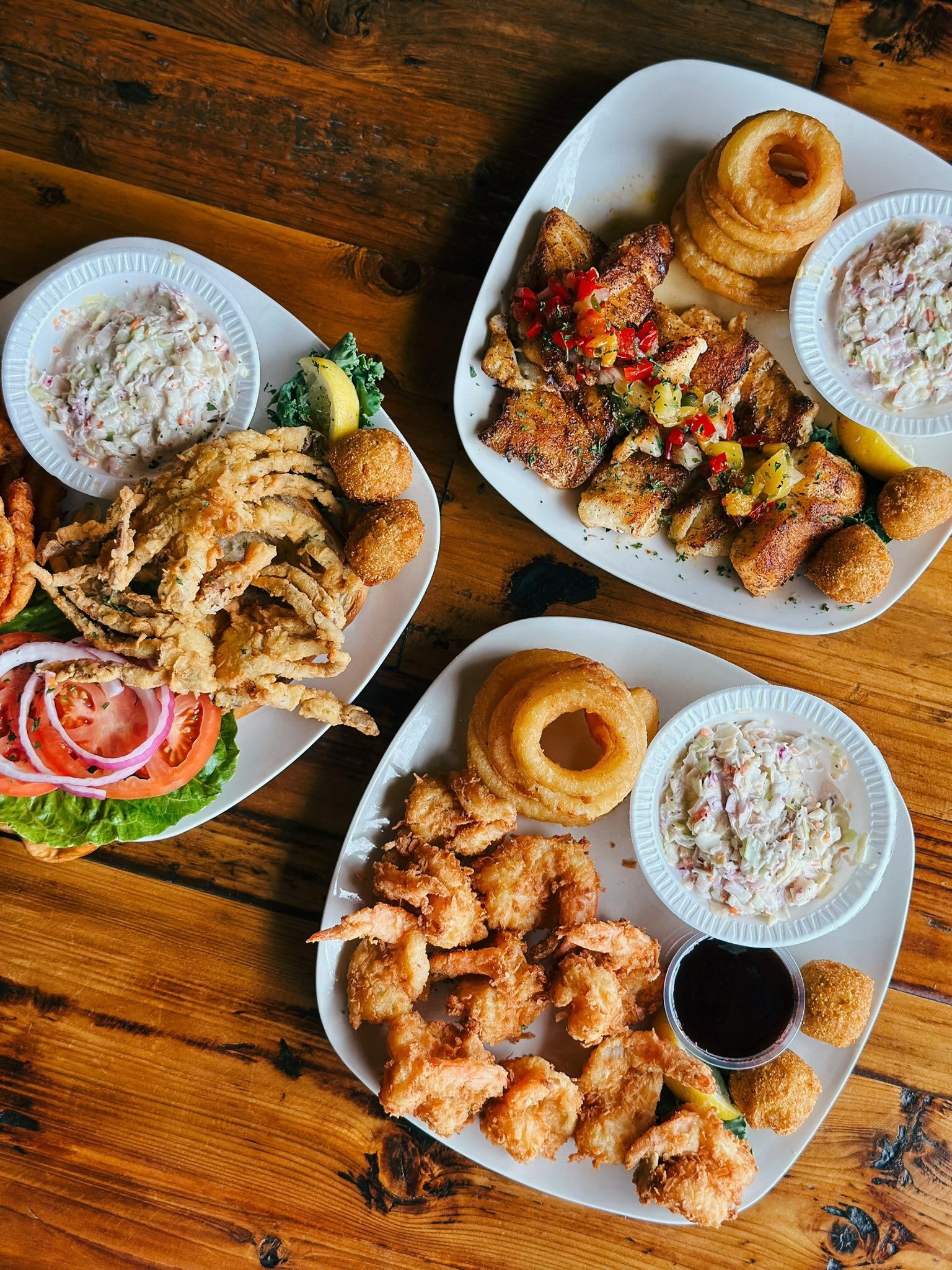 A wooden table with three plates of fried seafood, including shrimp and calamari, accompanied by lemon wedges and salad.