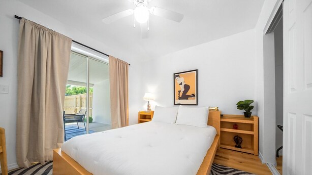 A bedroom with a large bed, a ceiling fan, a sliding glass door to a balcony, a bedside table with a lamp, and a small shelf with decorative items. - The Happy Haven Near Manatee Tours (Crystal River)