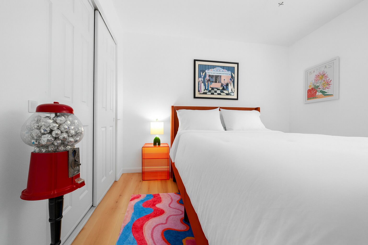 A hotel room with a bed, a nightstand with a lamp, a red gumball machine, and a colorful rug.