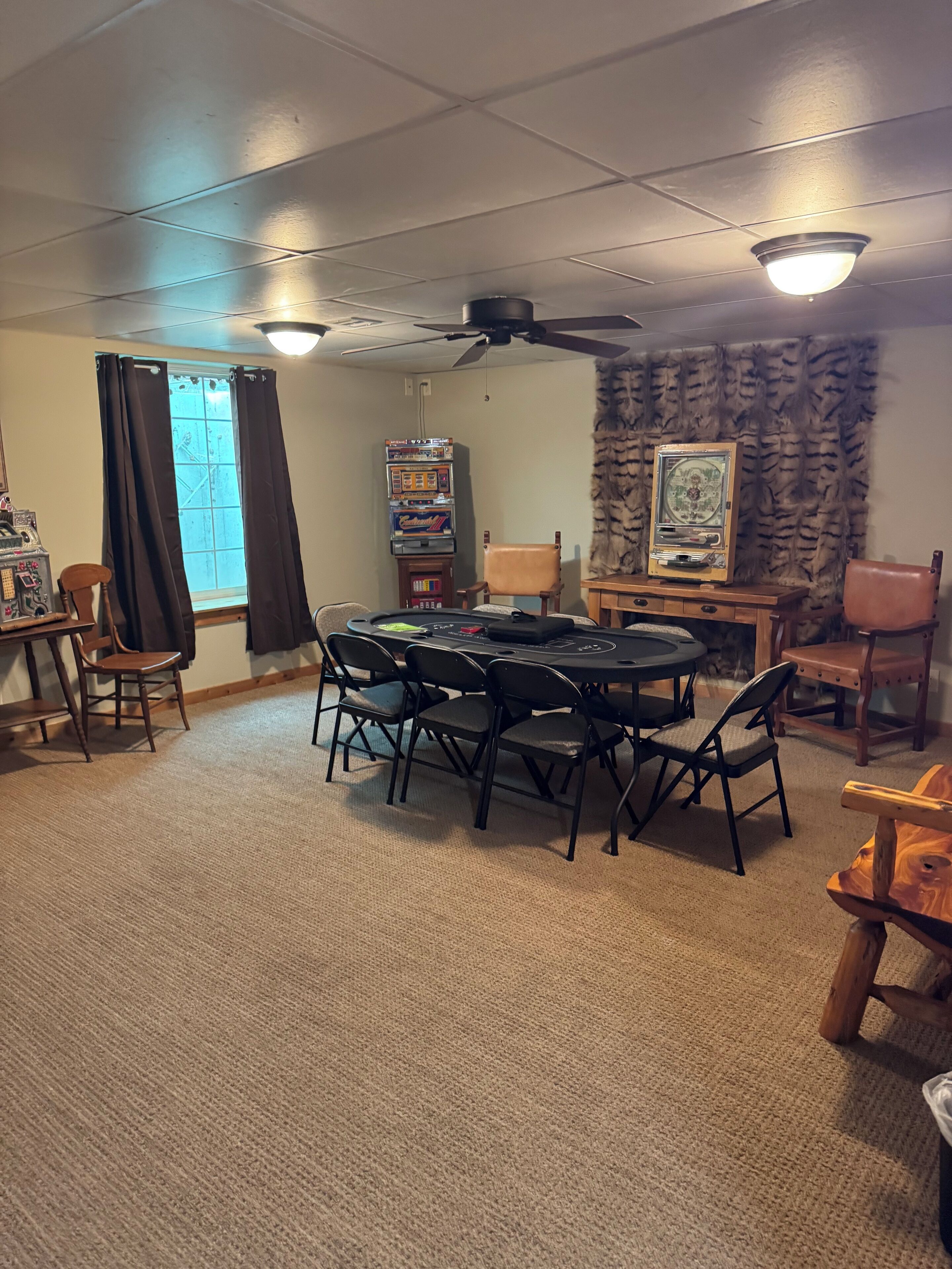 Une pièce comprenant une table ronde, des chaises, un ventilateur de plafond, une fenêtre avec des rideaux et un mur orné d’un panneau décoratif.
