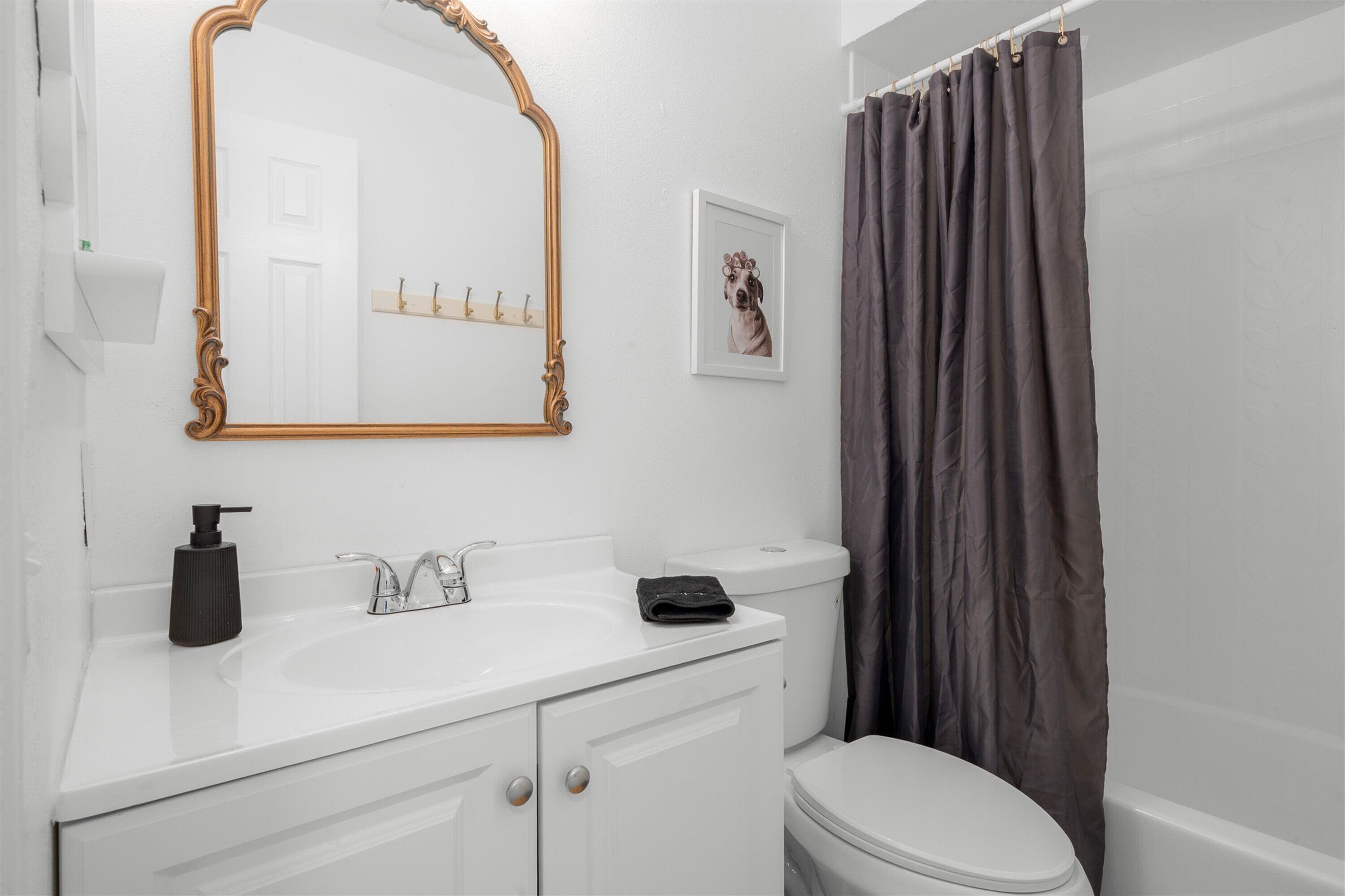 Une salle de bain avec un lavabo blanc, un miroir doté d’un cadre orné, des toilettes et un rideau de douche.