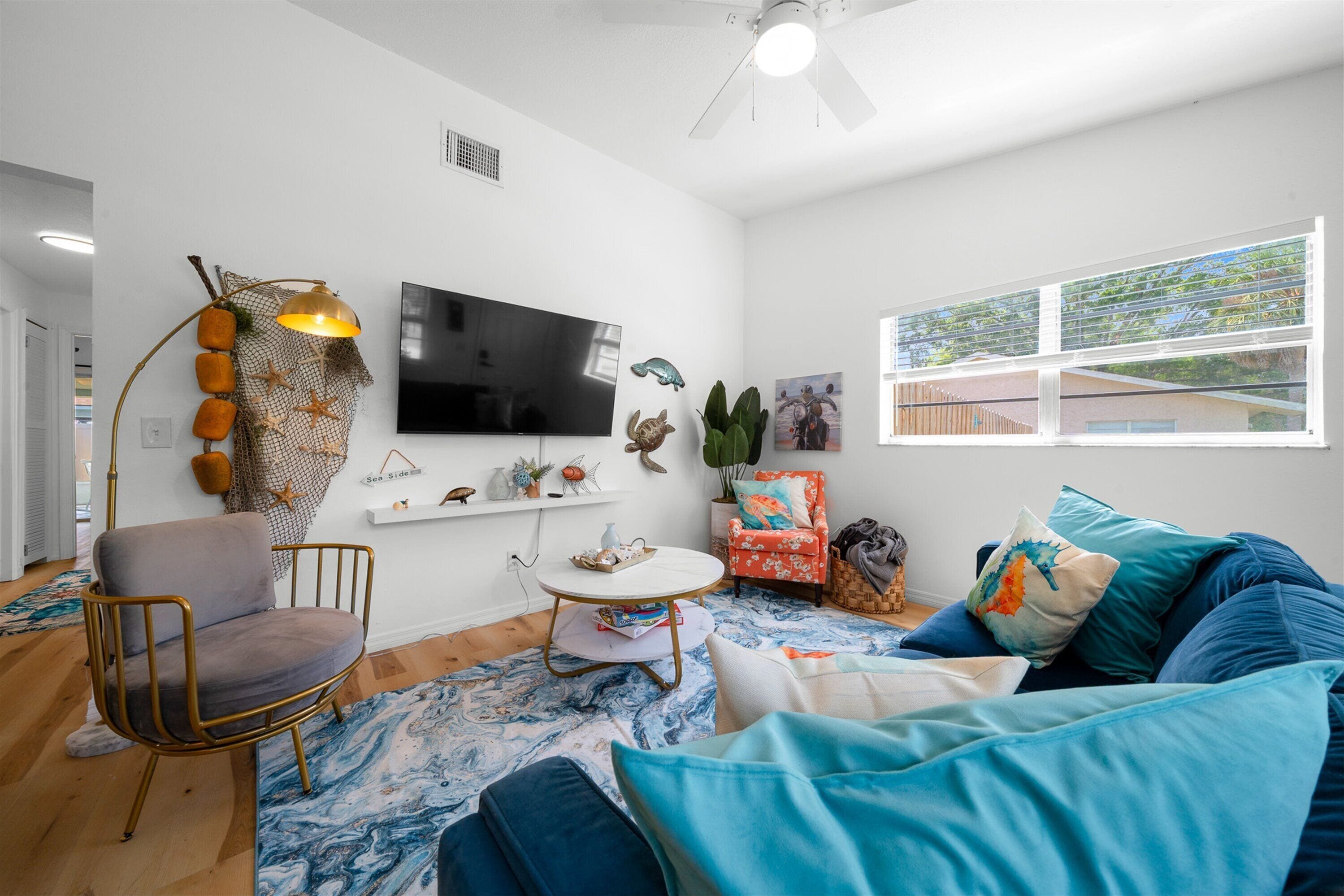 A modern living room with a flat-screen TV, a round coffee table, a comfortable armchair, a sofa with blue cushions, and a large window with a view.