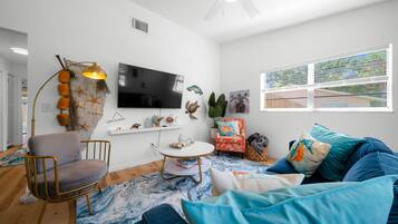 A modern living room with a flat-screen TV, a round coffee table, a comfortable armchair, a sofa with blue cushions, and a large window with a view.