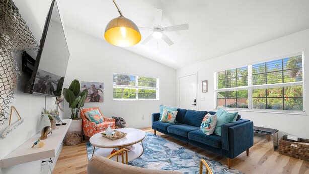 Ein Wohnzimmer mit einem blauen Sofa, einem runden Couchtisch, einem Deckenventilator und großen Fenstern mit Grünpflanzen draußen.