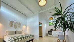 Quarto com cama, mesas de cabeceira, um painel de macramé na parede, kitchenette e um vaso com planta.