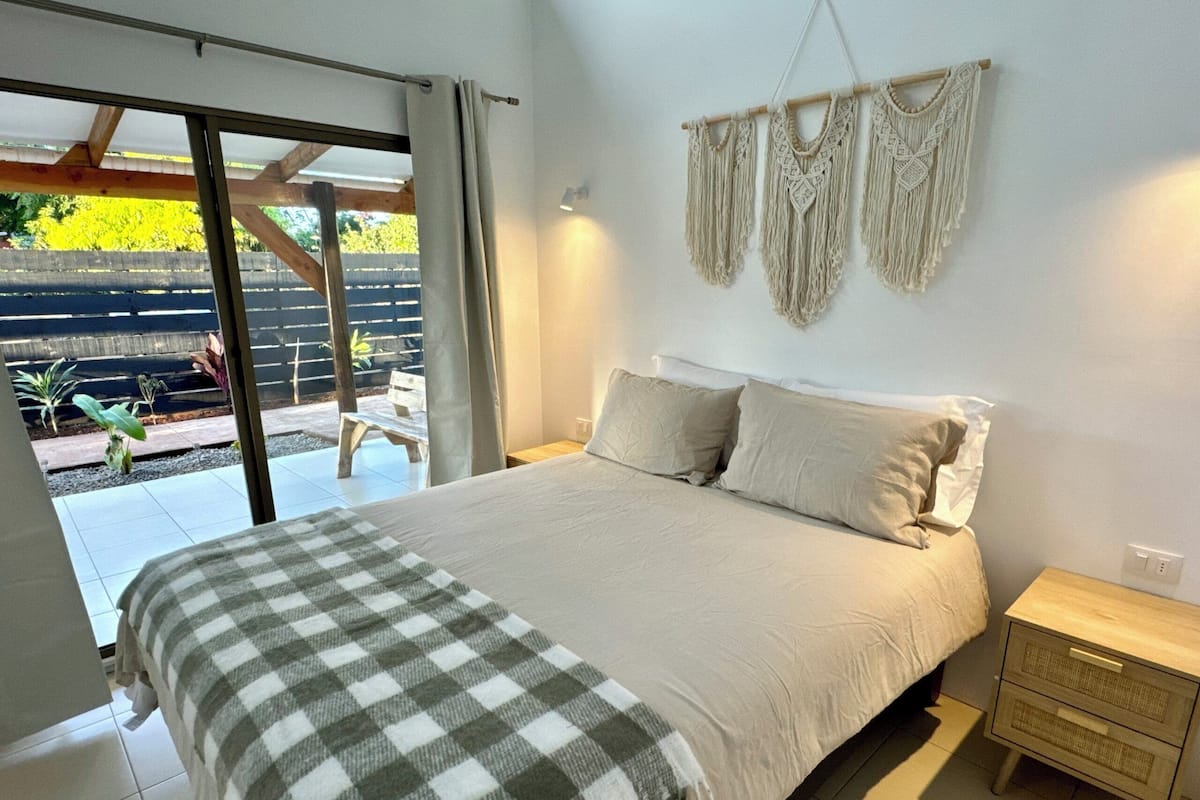 Une chambre à coucher avec un lit, une table de chevet, une décoration murale en macramé et une vue sur l’extérieur.