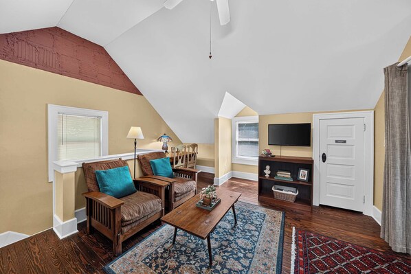 A cozy living room with a sloped ceiling, wooden furniture, a flat-screen TV, and a door with a nameplate.