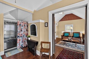 A room with a wooden floor, a black chair, a wooden table, a sofa with teal cushions, a window with blinds, and a door leading to another room.