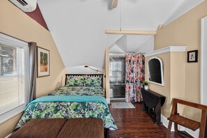 A bedroom with a bed, a desk, a chair, a window with blinds, and a door leading to a bathroom. - Historic Studebaker Carriage House- Upper Level (South Bend)