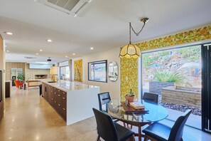 A modern kitchen with a central island, a dining area with a glass table, and a view of a landscaped garden through large windows. - Paradise Valley Private Hilltop Estate--Entertainers Dream (Paradise Valley)