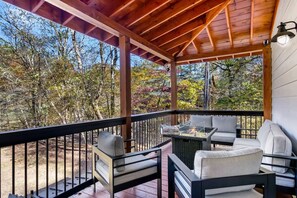 A covered outdoor patio with a fire pit table and two sofas, surrounded by a wooden railing and overlooking a forested area. - Riverfront Cabin, GameRoom, Fenced, 10 min to town (Ellijay)