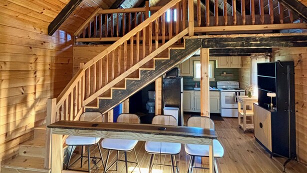 A rustic wooden interior with a staircase leading to an upper level, a kitchen area, and a bar with stools. - Charming 1 BR Cabin at Persimmon & Pine Hollow- Secluded Feel, Close to Town (Dublin)