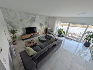 Uma sala de estar moderna com uma televisão de tela plana grande, um sofá cinza e duas plantas em vasos.