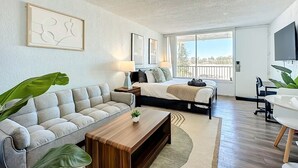 A modern hotel room with a sofa, a wooden coffee table, a bed with a view, a TV, and a balcony. - 5-202 Breakwater (Jacksonville)