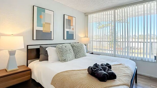 A neatly made bed with a patterned throw, two bedside lamps, and abstract artwork on the wall. - 5-202 Breakwater (Jacksonville)