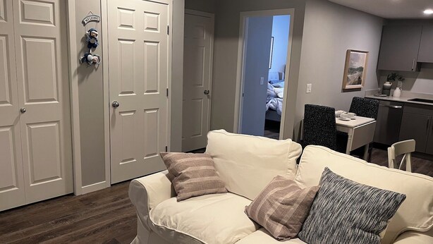 A living room with a beige sofa, two pillows, and a small rug. There is a dining area with a table and chairs, and a kitchen in the background. - Modern 1BD Suite•5 star Comfort•Beaumont•Edmonton (Beaumont)