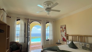 Um quarto com cama, poltrona, ventilador de teto e vista para o mar.