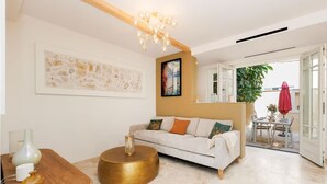 Una sala moderna con un sofá blanco, una mesa de centro redonda dorada y una mesita auxiliar de madera. En la pared hay un cuadro y se distingue un comedor con mesa y sillas a través de una puerta abierta.