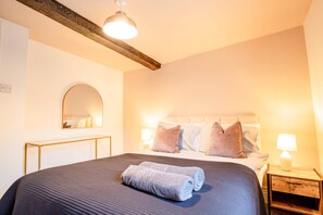 Un letto rifatto con cura, con copriletto scuro e due asciugamani arrotolati, un comodino con una lampada e uno specchio appeso al muro.