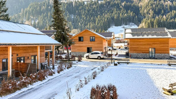 Une cabane en bois avec un porche couvert, une voiture garée devant, et un paysage enneigé avec des montagnes en arrière-plan.