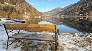 Un banc en bois au bord d’un lac paisible, la neige recouvrant le sol, entouré de montagnes boisées.