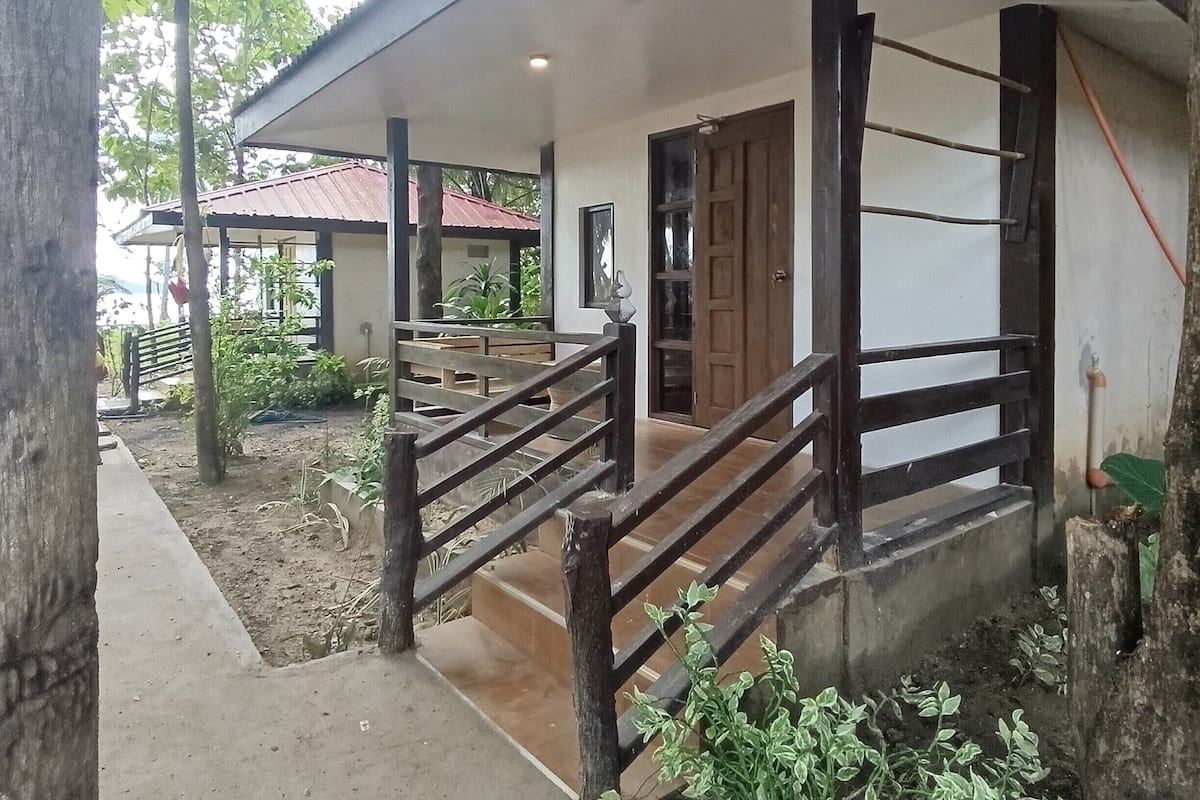 A wooden porch with a metal railing leading to a door, surrounded by greenery and a concrete path.