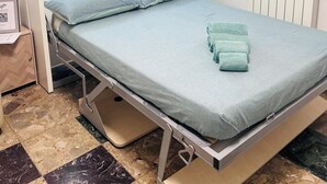 Une petite chambre comprenant un lit, une petite table de chevet, une fenêtre avec des rideaux légers et un radiateur.