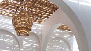 Une pièce baignée de lumière naturelle, avec deux chaises longues en bois, un luminaire suspendu en fibres tressées et une grande fenêtre donnant sur un paysage verdoyant.