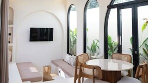 Un salon moderne avec une table à manger ronde, des sièges confortables, un ventilateur de plafond et de grandes fenêtres ornées de plantes.