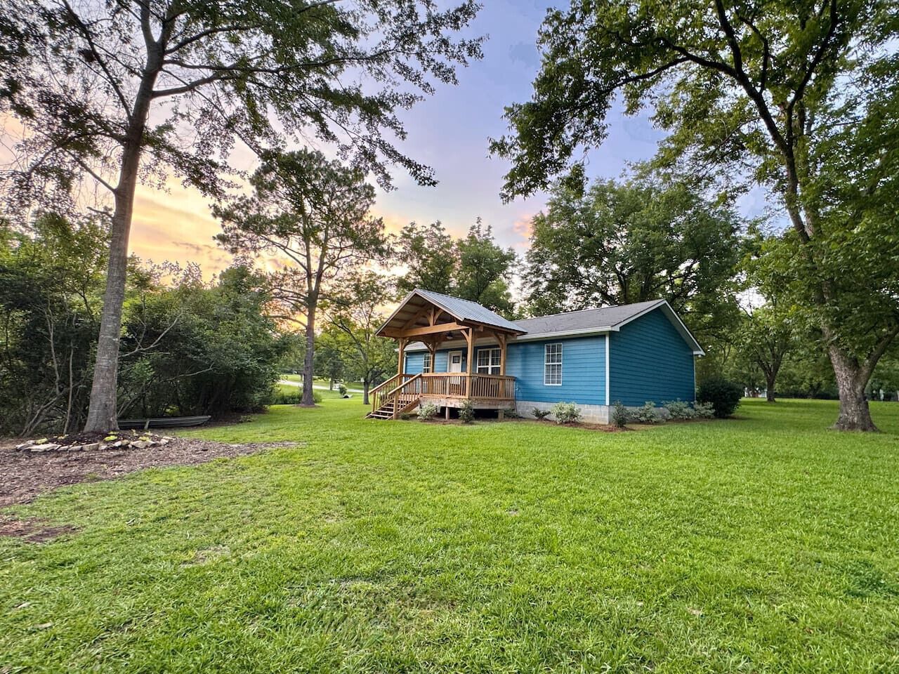 A single-story blue house with a wooden porch and a small deck, surrounded by a well-maintained lawn and trees.