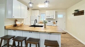 Une cuisine avec des meubles blancs, un îlot central en bois doté de tabourets et des luminaires suspendus.