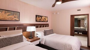 Habitación de hotel con dos camas, una mesita de noche con lámpara, obras de arte en las paredes y un baño visible a través de una puerta abierta.