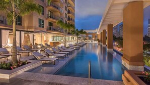 A hotel pool area with sun loungers and a view of the city skyline at dusk. - Waikiki 1BR at Grand Islander – Full Kitchen, Beach & Resort Fun at Hilton Villa (Honolulu)