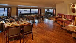 A restaurant with wooden flooring, multiple dining tables, and chairs. Large windows offer a view of the ocean. - Waikiki 1BR at Grand Islander – Full Kitchen, Beach & Resort Fun at Hilton Villa (Honolulu)