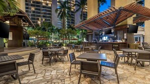 An outdoor patio area with stone flooring, wooden tables and chairs, and a bar area with a TV screen. - Waikiki 1BR at Grand Islander – Full Kitchen, Beach & Resort Fun at Hilton Villa (Honolulu)