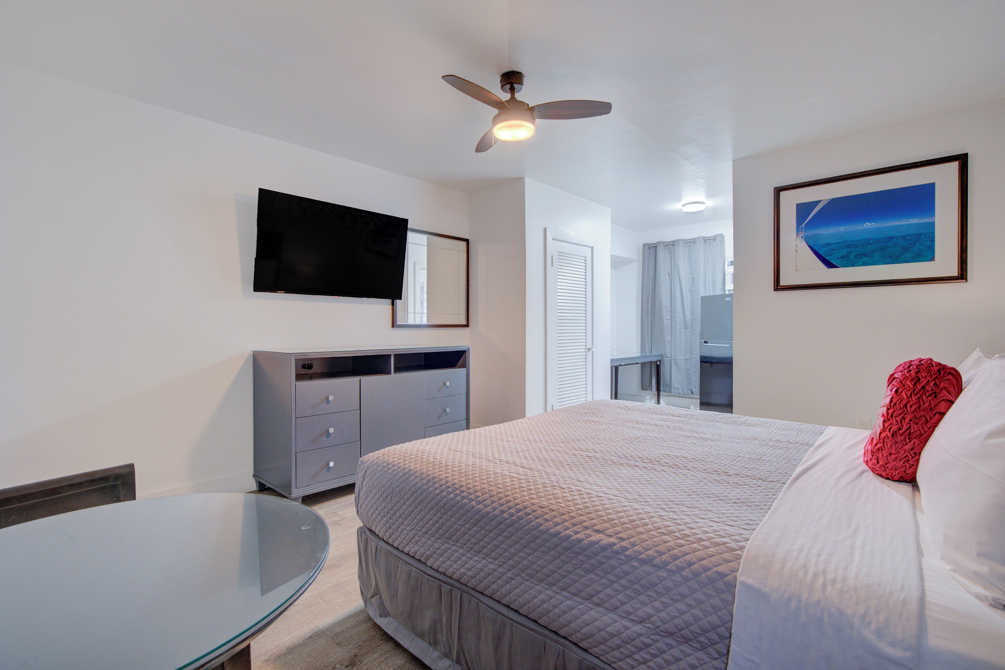 A hotel room with a bed, a desk, a ceiling fan, a TV mounted on the wall, and a framed picture on the wall.