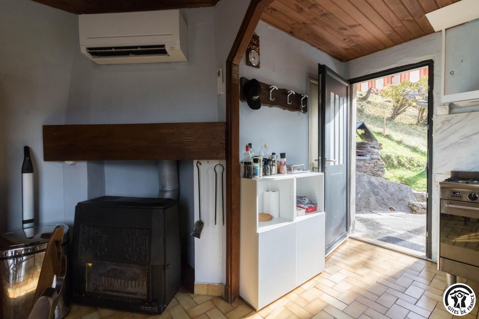 Une pièce chaleureuse avec un poêle à bois, une étagère remplie d’ustensiles de cuisine et une vue sur un mur de pierres à l’extérieur.