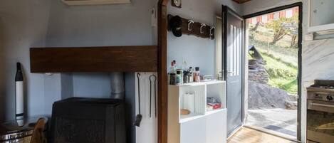 Une pièce chaleureuse avec un poêle à bois, une étagère remplie d’ustensiles de cuisine et une vue sur un mur de pierres à l’extérieur.