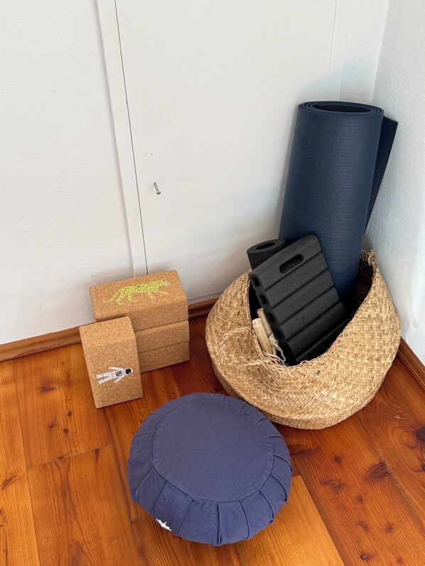 A corner of a room with a wooden floor, a woven basket containing black foam rollers, a rolled-up yoga mat, and two cork storage boxes with animal designs.