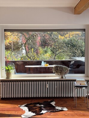 Ein Zimmer mit einem großen Fenster, einem Heizkörper, einem Kuhfellteppich, einem kleinen Tisch mit einer Pflanze und einem Metallstuhl.
