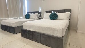 Due letti con poggiatesta grigi e biancheria da letto bianca, un comodino con una lampada e una finestra con tende leggere.