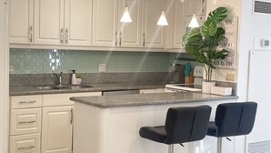 Una cucina moderna con mobili bianchi, un piano di lavoro in granito, due sgabelli da bar neri e illuminazione a sospensione.