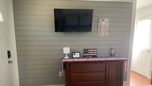 Un dormitorio con cama, cómoda, televisor montado en la pared y una mesita de noche con lámpara.