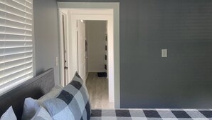 Une chambre avec un sol à carreaux, un lit avec des oreillers, une porte donnant sur un couloir et un interrupteur sur le mur.