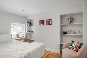 Une chambre à coucher avec un grand lit, une chaise, une étagère ornée d’objets décoratifs et une fenêtre avec des stores.