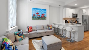 A modern living room with a grey sofa, colorful cushions, a painting of red flowers, a kitchen with white cabinets, and a bar area with stools. - Spacious 4BR Rittenhouse Square Retreat (Philadelphia)