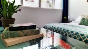 Une chambre à coucher avec un lit, une fenêtre donnant sur des immeubles, une plante en pot et une table avec des livres.