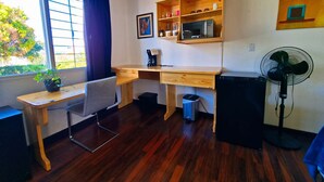 Habitación compacta con un escritorio de madera, una silla, un cesto de basura pequeño, un altavoz negro y un ventilador de pie.